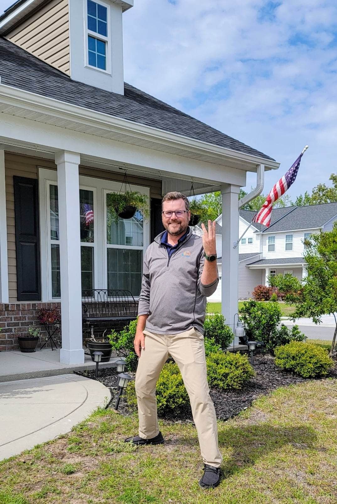 Man in front of a house, holding up three fingers. Blue sky, American flag.