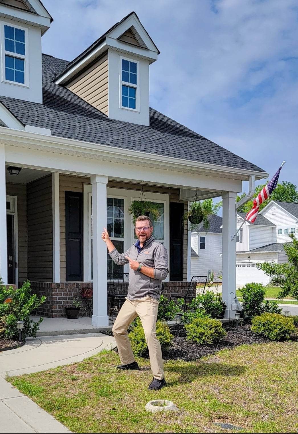 Man poses in front of a house, mimicking a karate chop. House has a porch, American flag. Sunny day.