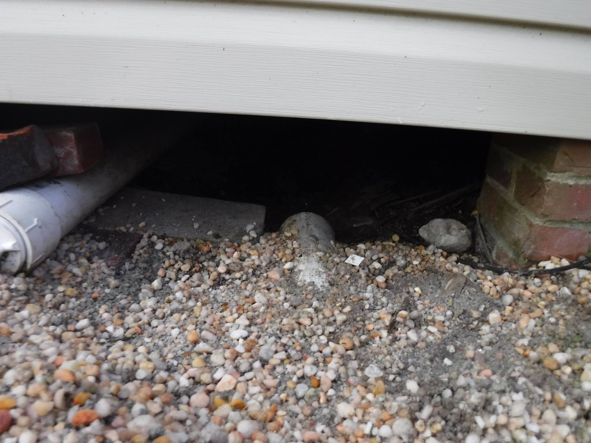 Gravel-covered crawlspace under a building with pipes, brick and a dark interior.