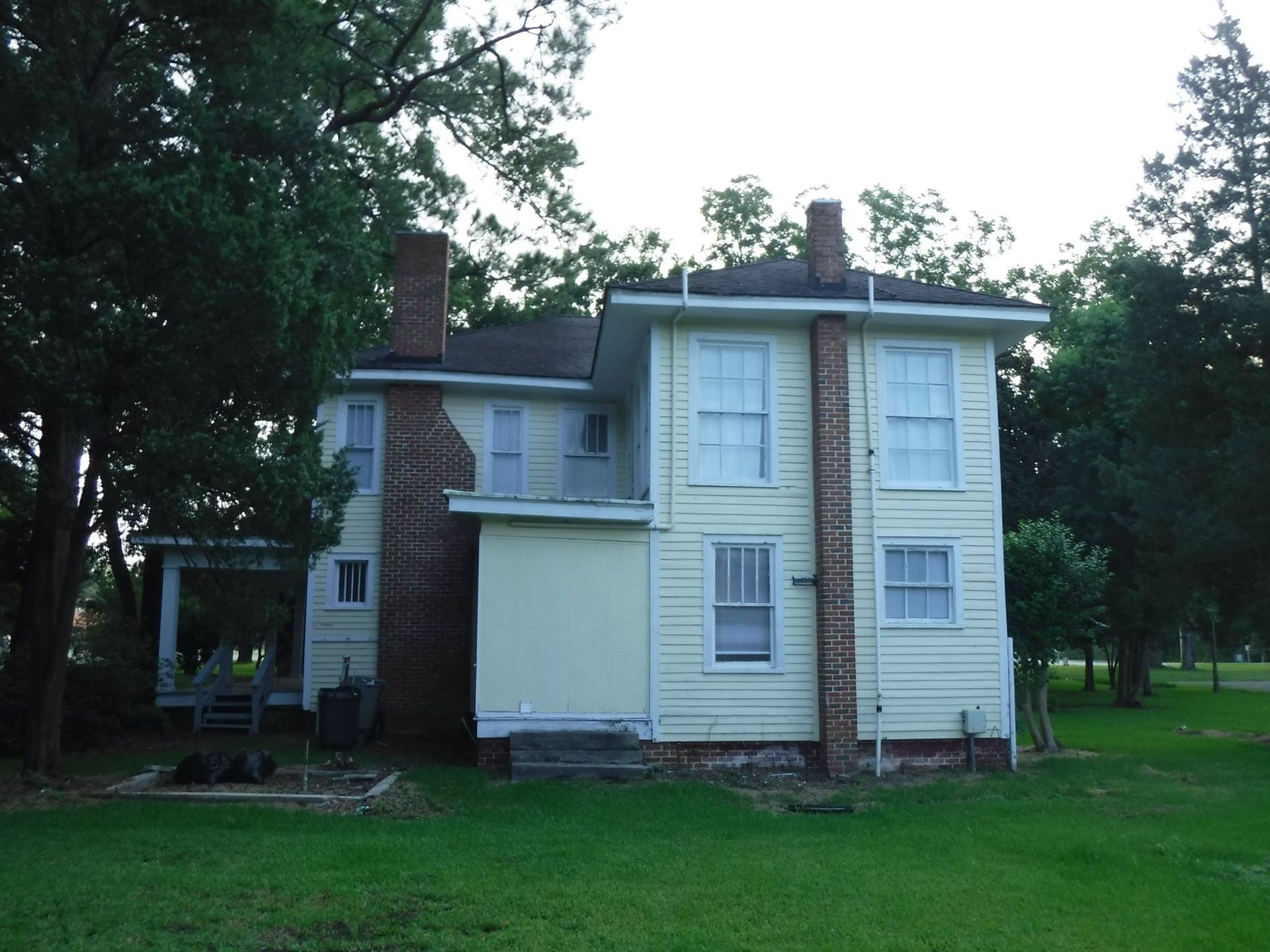 Two-story, light yellow house with brick chimneys and white window frames, surrounded by trees and green grass.