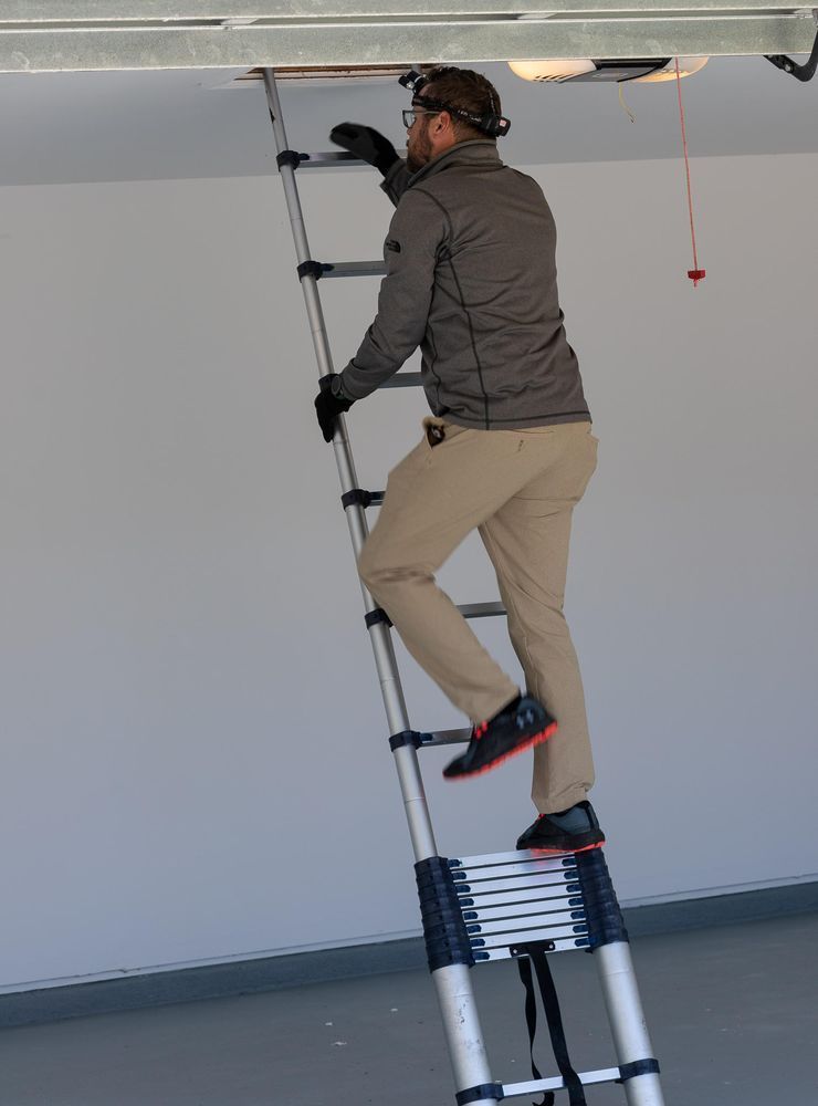 Person on a telescoping ladder reaching into a ceiling. They wear gloves, a jacket, and pants. Indoors.