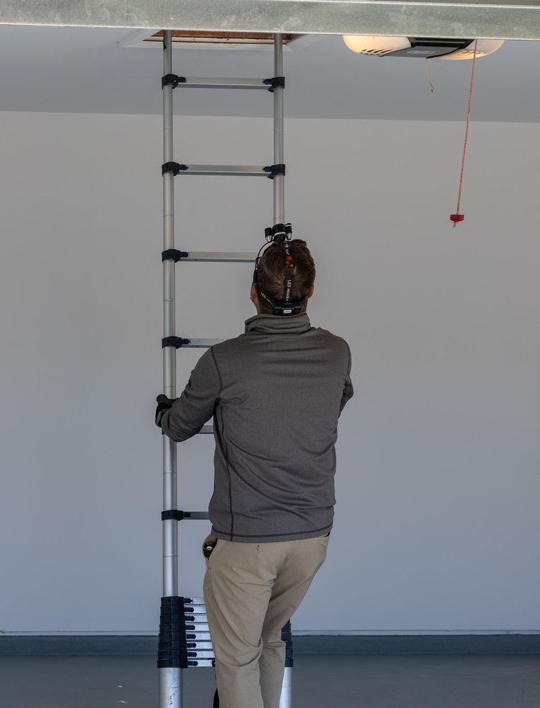Person climbing a silver telescopic ladder to a ceiling opening in a white room.