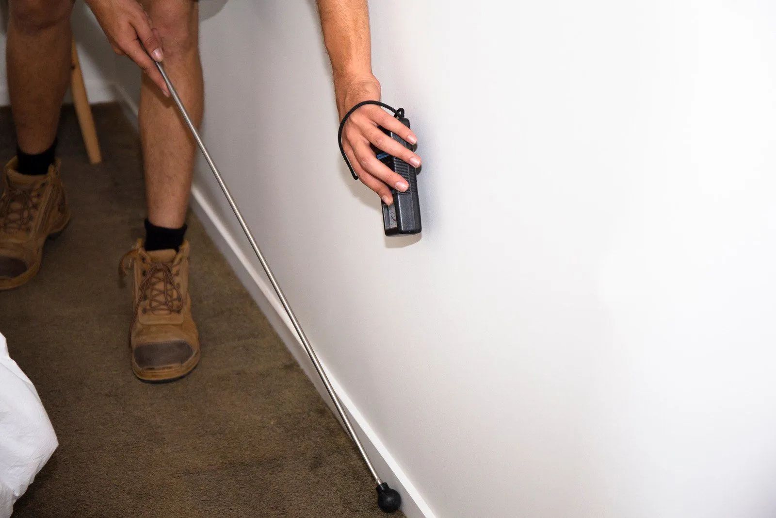 A person uses a telescopic inspection mirror to check behind an object against a white wall in a room with carpet.