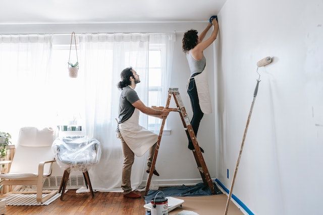 Two people painting a wall, surrounded by supplies. One person stands on a ladder while the other person holds it.