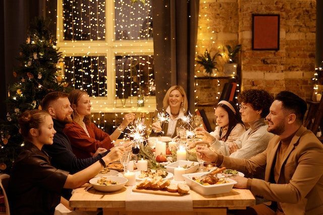 Family sitting at a rectangular wooden table served with cups and plates of food; there's a window covered in twinkle lights in the background and, next to it, a Christmas tree