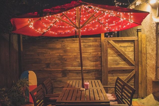 Red patio umbrella lined with string lights, open over a brown wooden table and four matching chairs