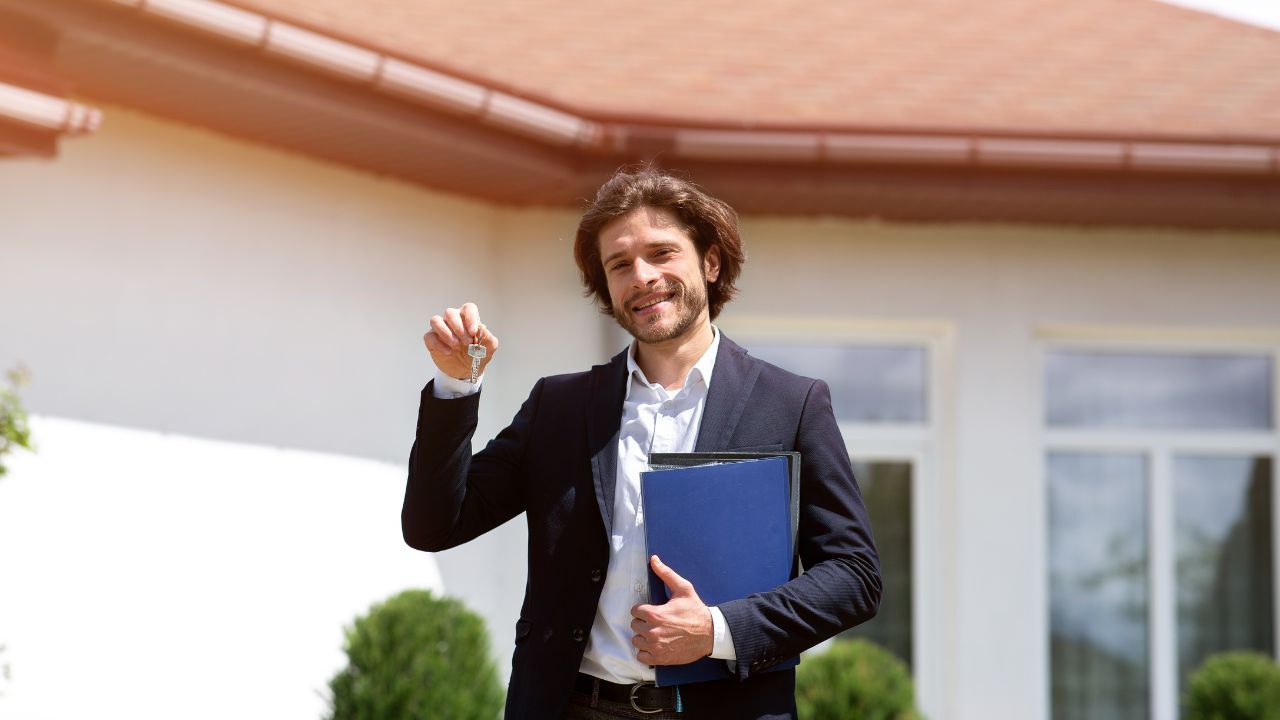 Smiling real estate investor holding up a key in front of a house