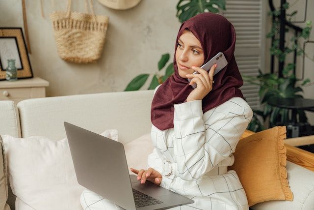 Person wearing a hijab, sitting on a couch, scrolling through their computer, and making a phone call