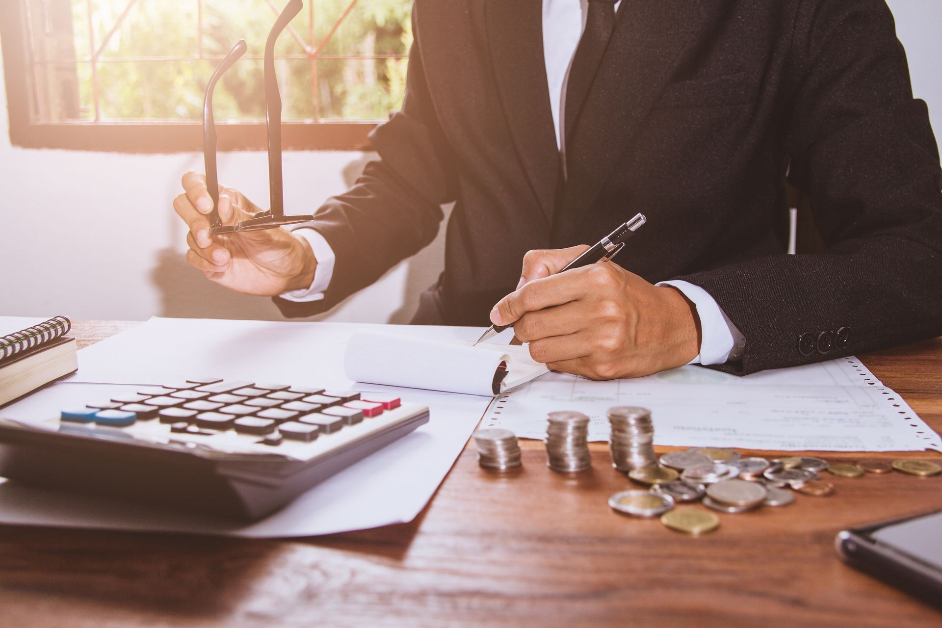 A person in a business suit writing on a document at a desk with a calculator and piles of coins nearby.