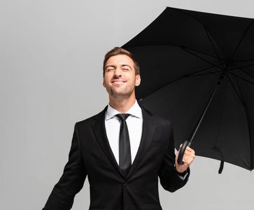 A smiling person in a suit holds an open black umbrella against a light gray background.