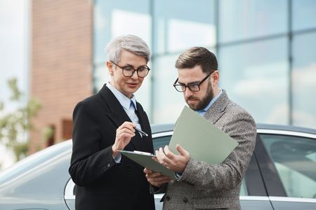 Two professionals in business attire stand outside by a car, looking over documents in a folder.