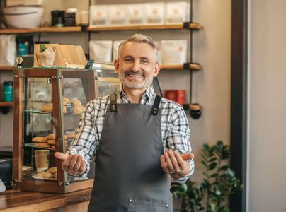 A smiling cafe worker in a plaid shirt and gray apron stands behind a counter with a display case of pastries.