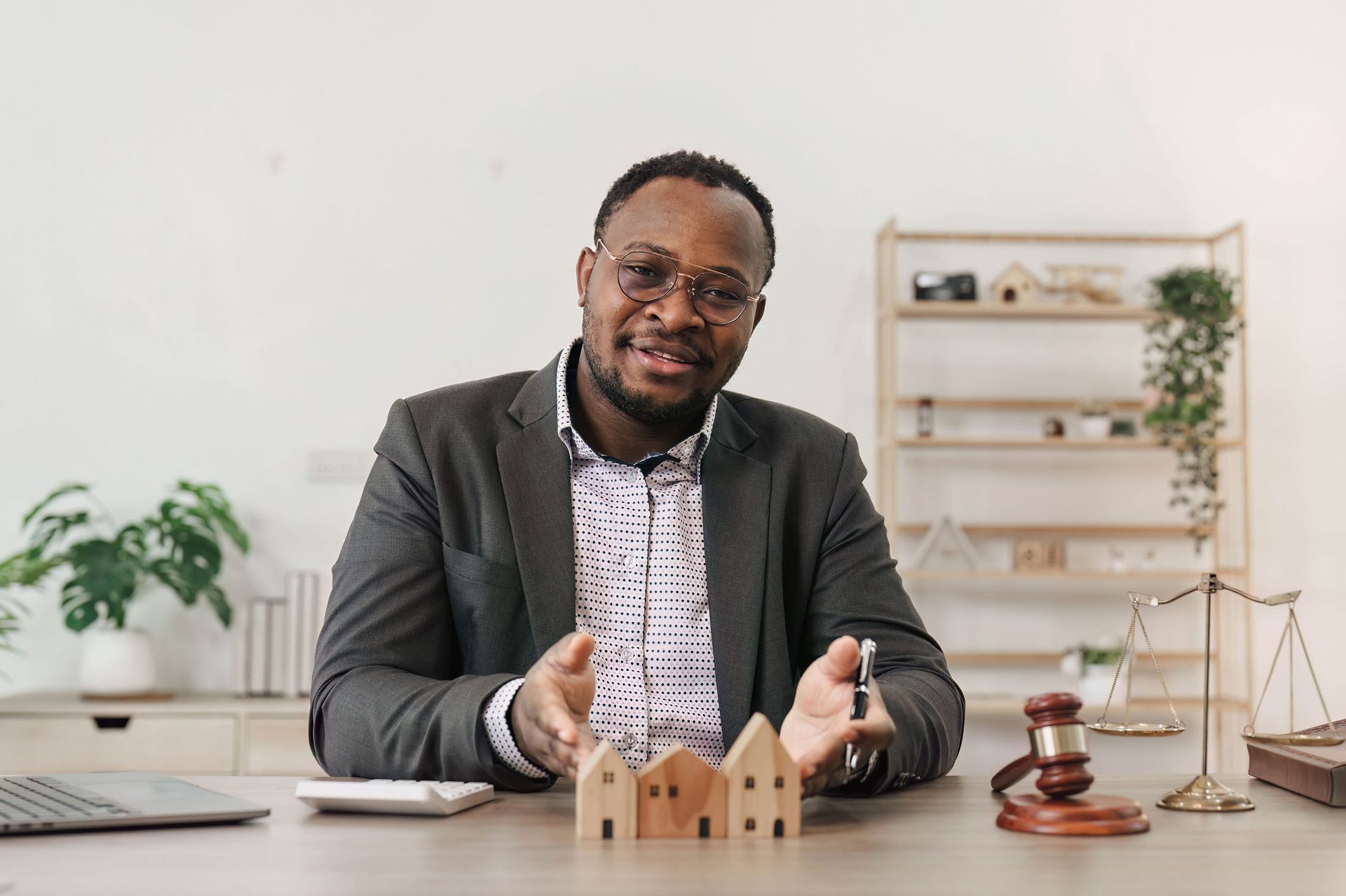 A professional sitting at a desk with wooden house models, a gavel, and scales of justice in a bright, modern office.