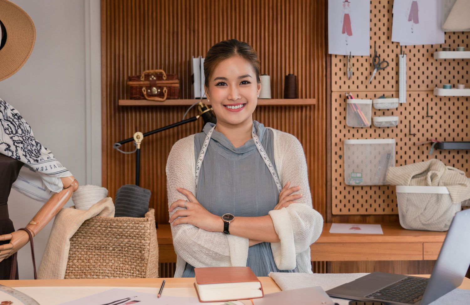 A smiling person with crossed arms in a workspace, standing before a wooden desk, a pegboard wall, and a sewing mannequin.