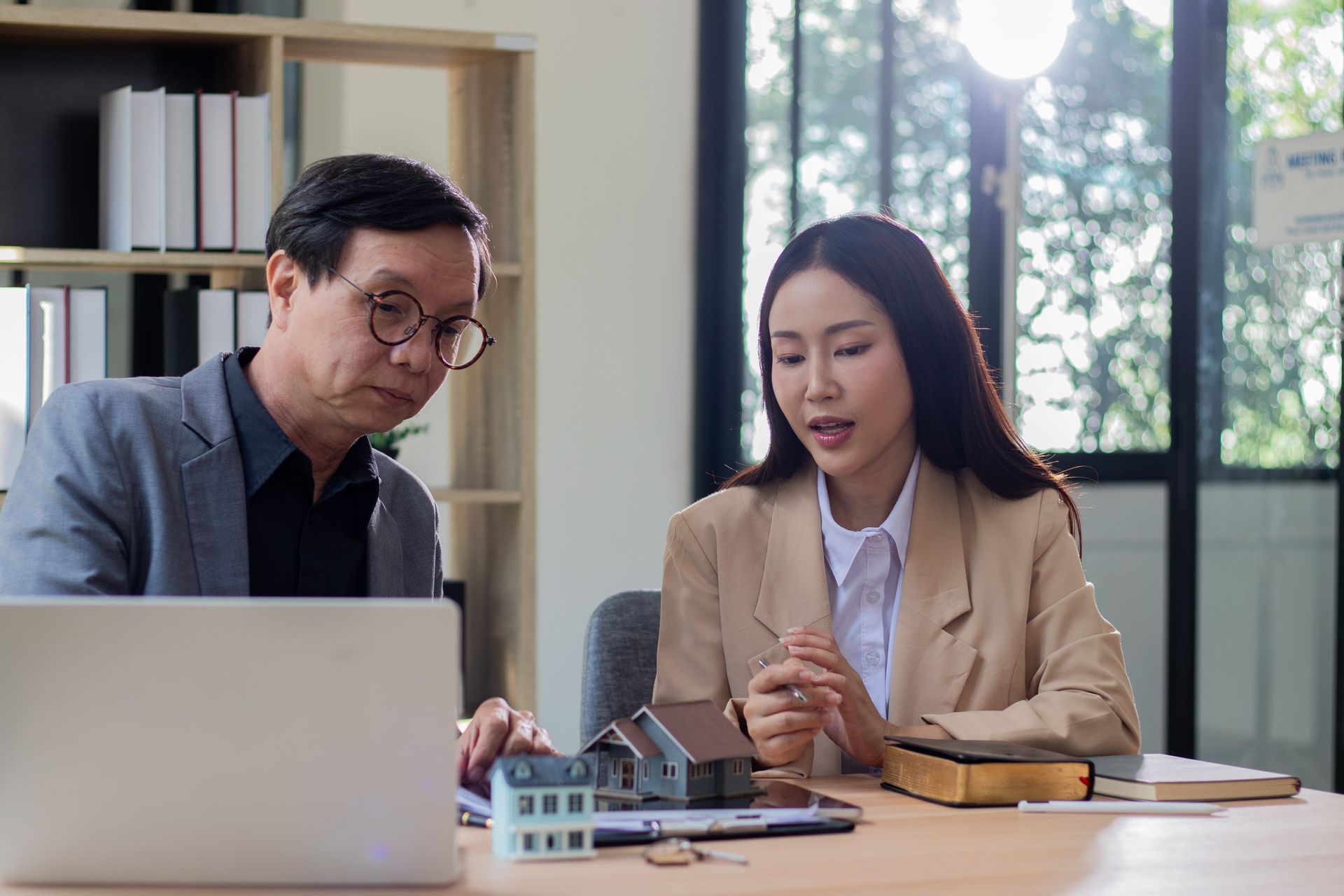 Two professionals at a desk, looking at a laptop and small house models while discussing a real estate project.