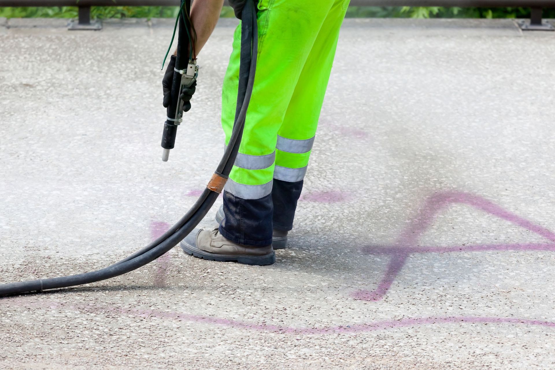 Worker in neon green pants using a pavement line-marking machine on asphalt.