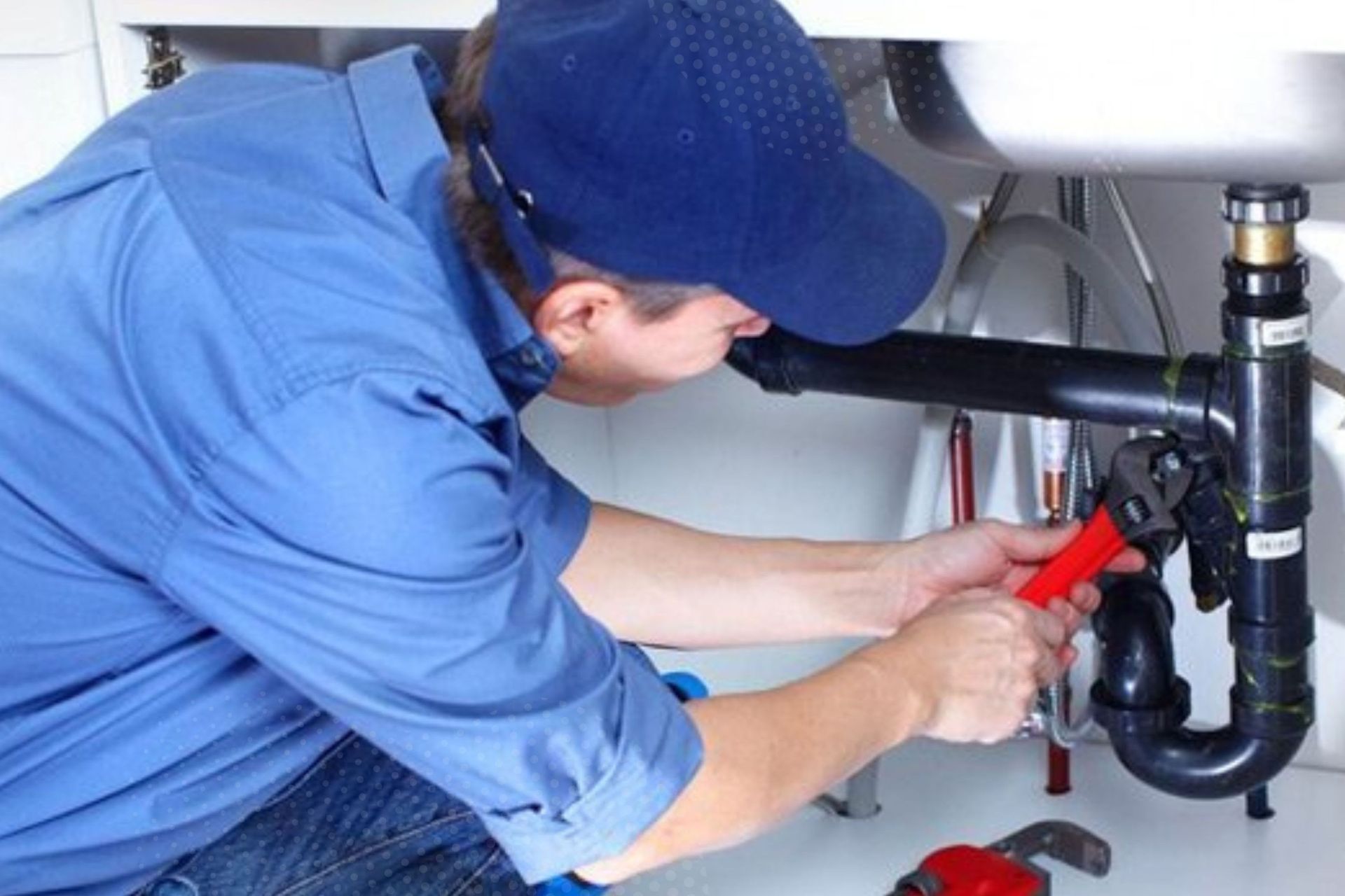 Plumber in blue uniform tightening black sink pipes under a bathroom sink with red-handled pliers