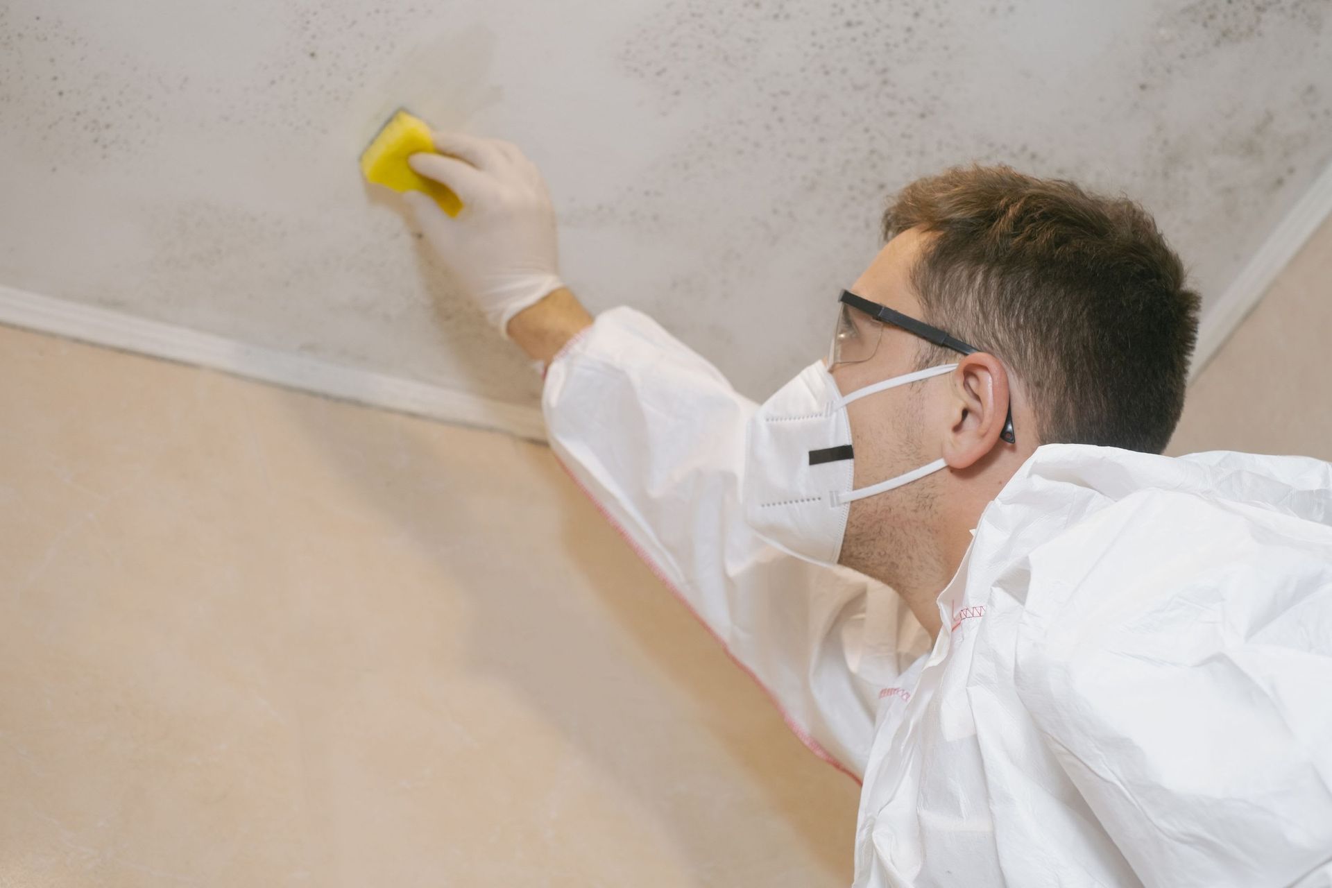 Person in protective gear cleaning a ceiling corner with a yellow sponge while wearing a mask and gloves