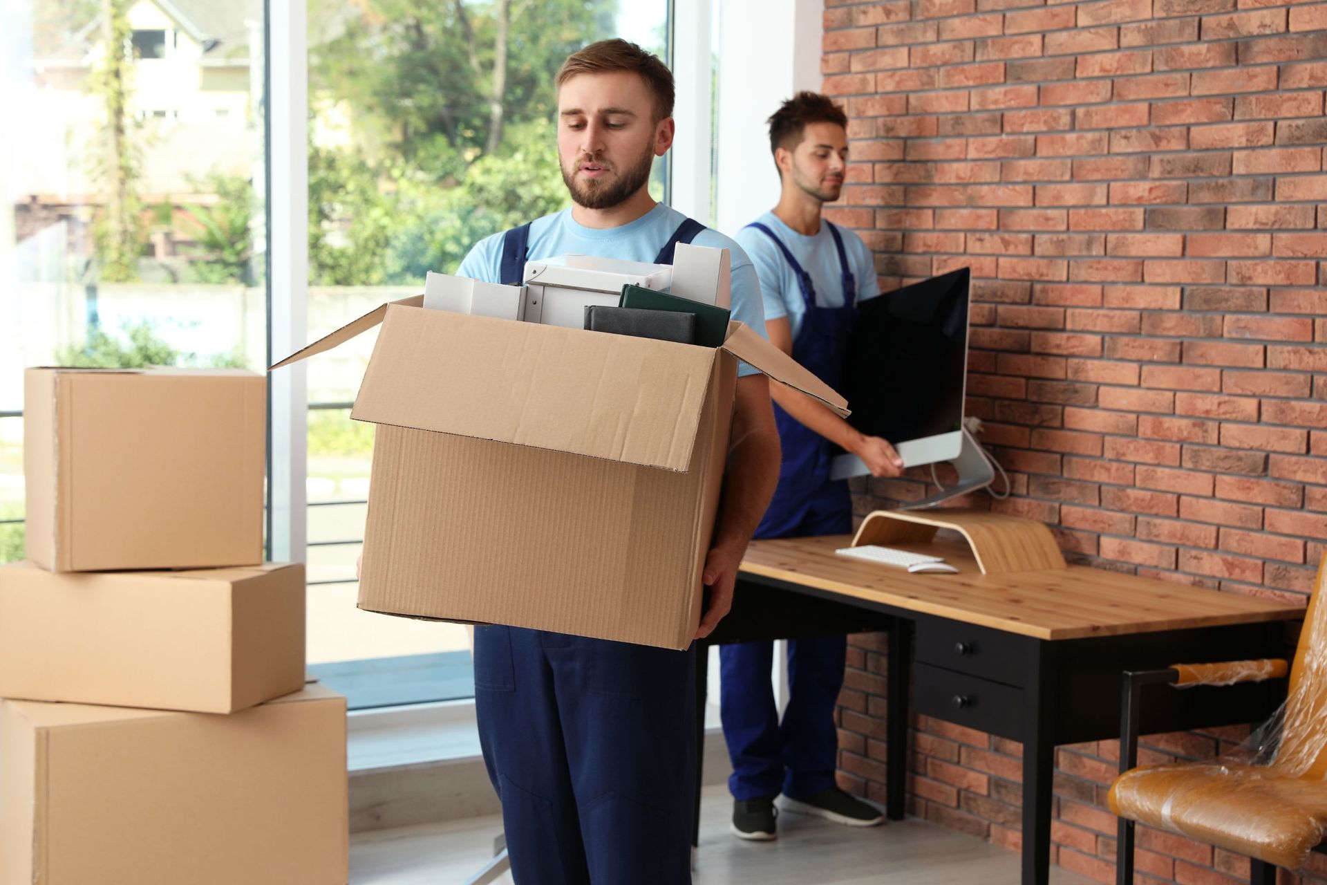 Two movers carrying boxes in a bright office with a desk and laptop.