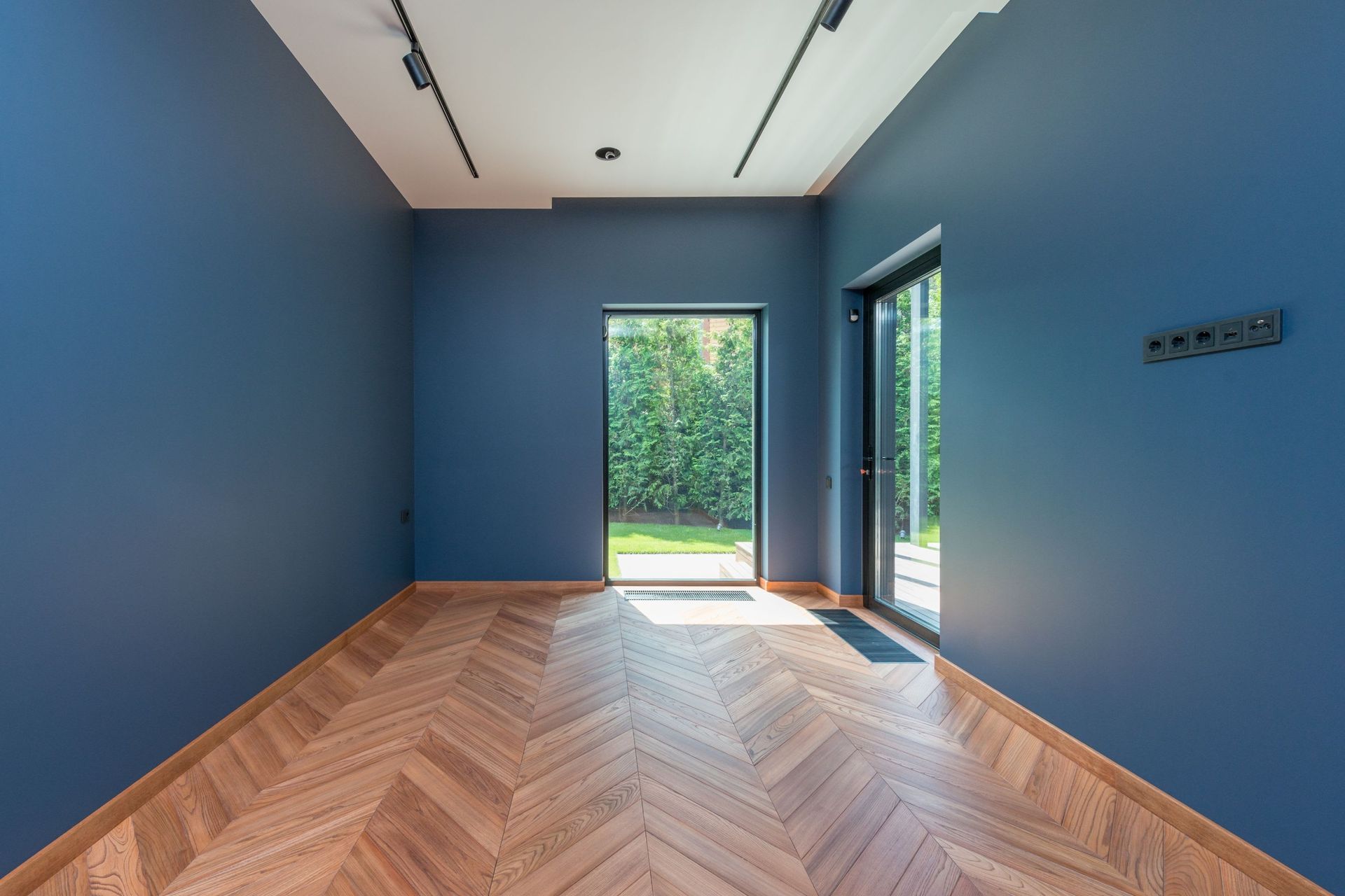 Empty blue room with herringbone wood floor and glass doors opening to greenery.