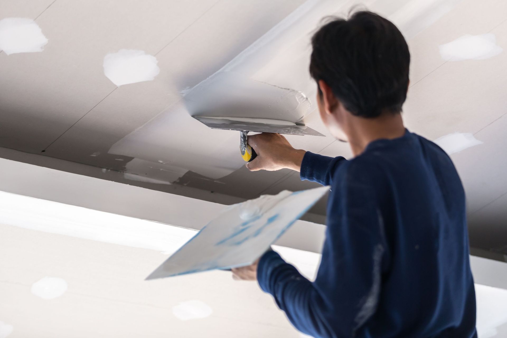 Person smoothing drywall with a putty knife while holding a drywall panel in a ceiling work area