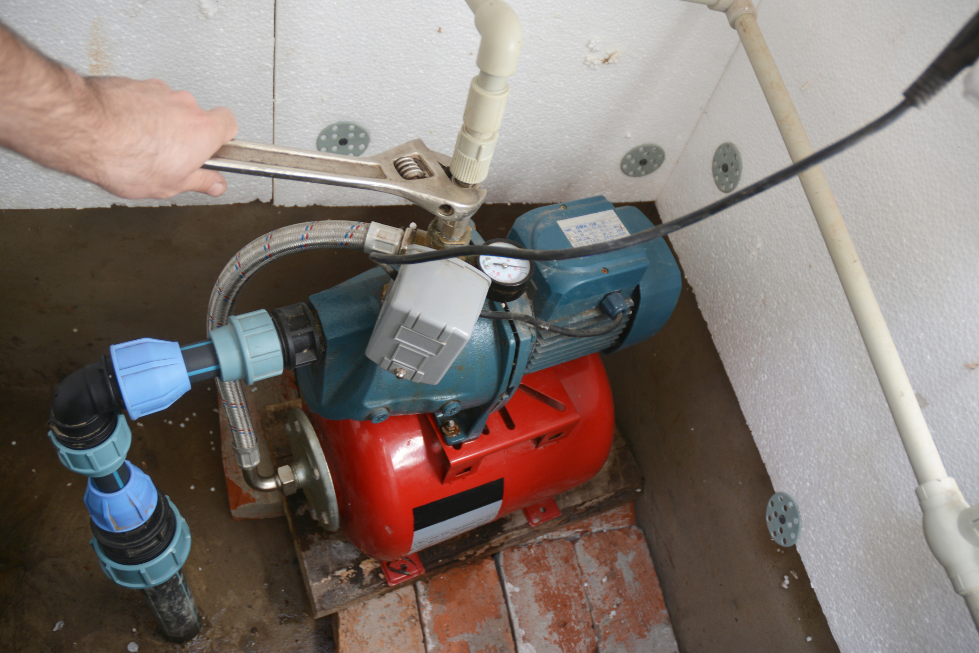 Blue and red water pump installed in a utility room, with pipes and a hand adjusting the valve