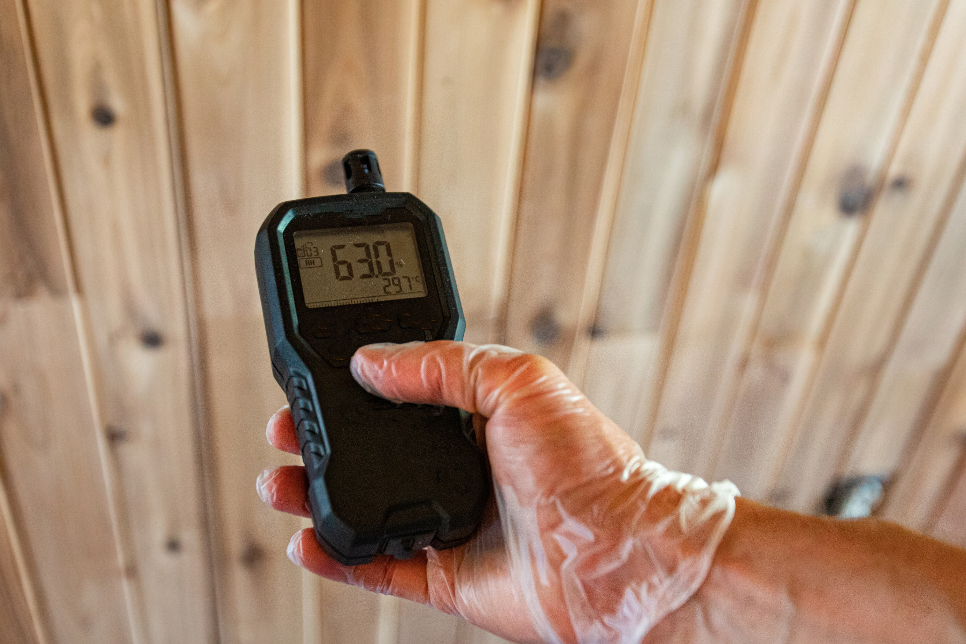Hand holding a digital meter in front of a wooden wall, showing a reading on the display