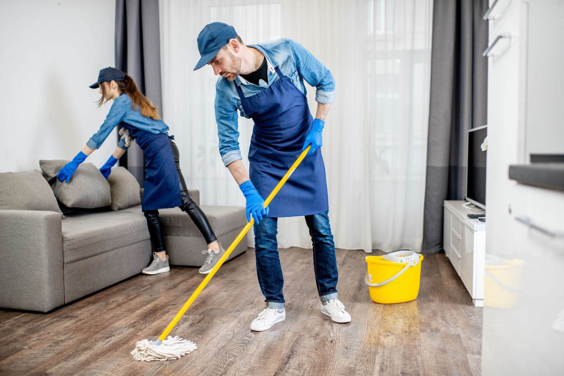 Two cleaners mopping a living room floor near a sofa and yellow bucket.