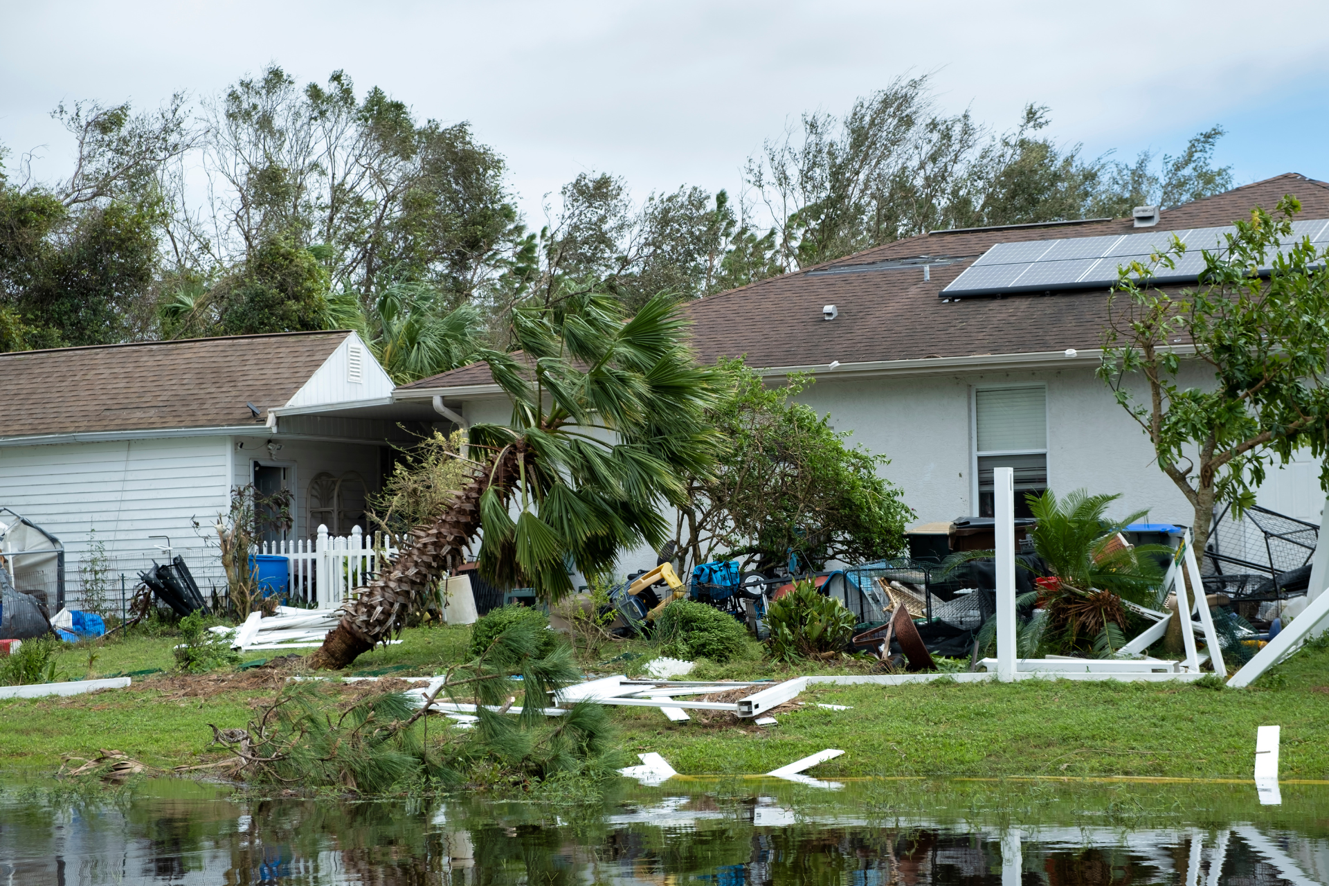 Storm-damaged suburban homes with debris, fallen trees, and standing water after severe weather
