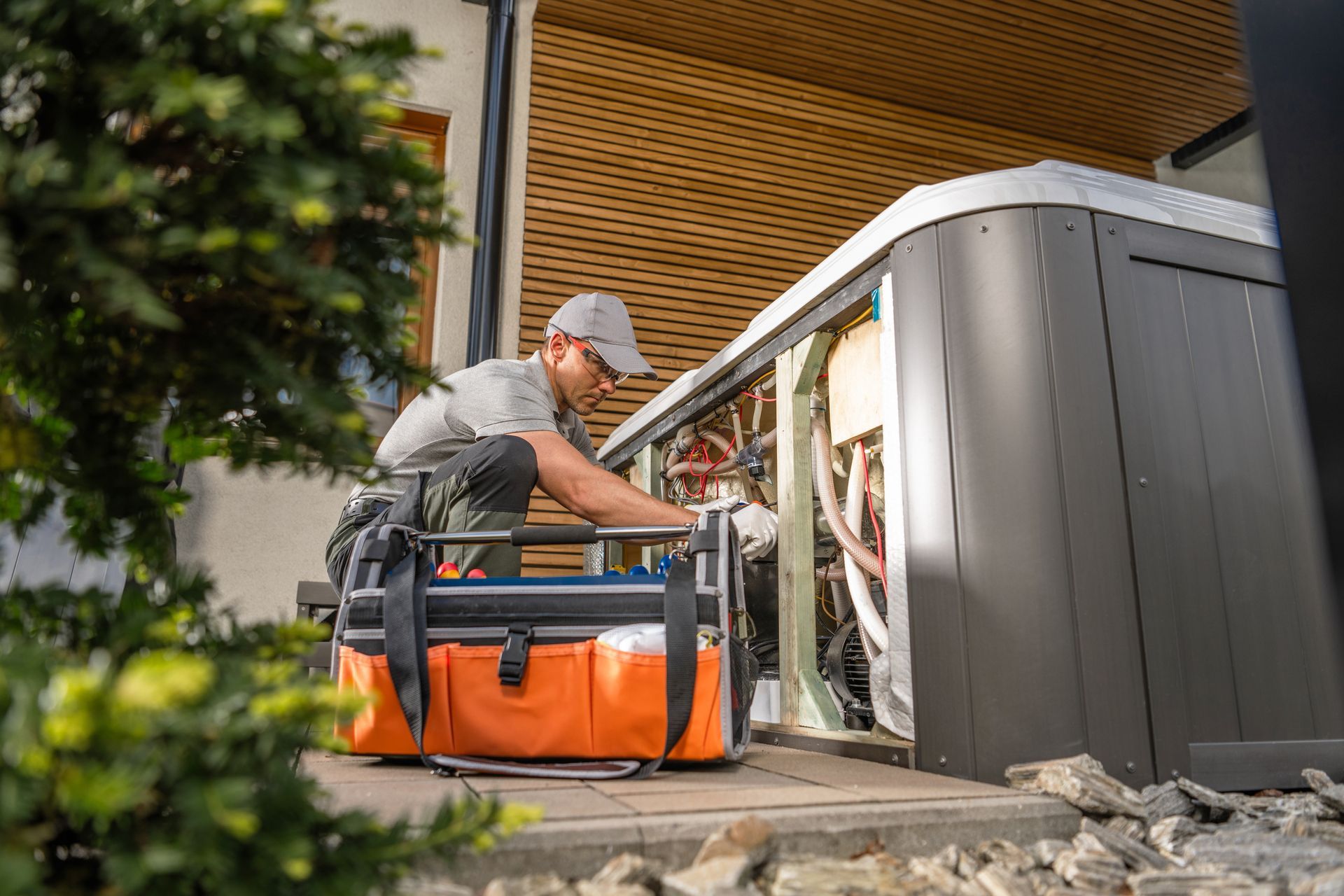 Technician working on outdoor HVAC unit beside a house, with tools and orange equipment bag.
