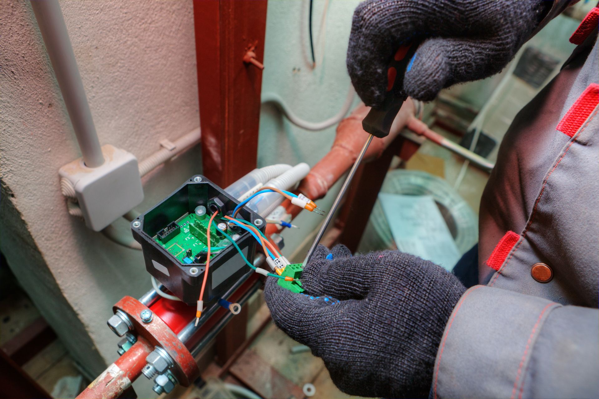 Gloved hands repair a pipe with wires and a circuit board in a tight utility space