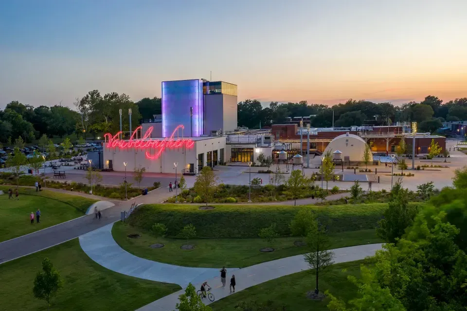Dusk view of a modern shopping center with a neon sign, lit tower, walking paths, and green lawn