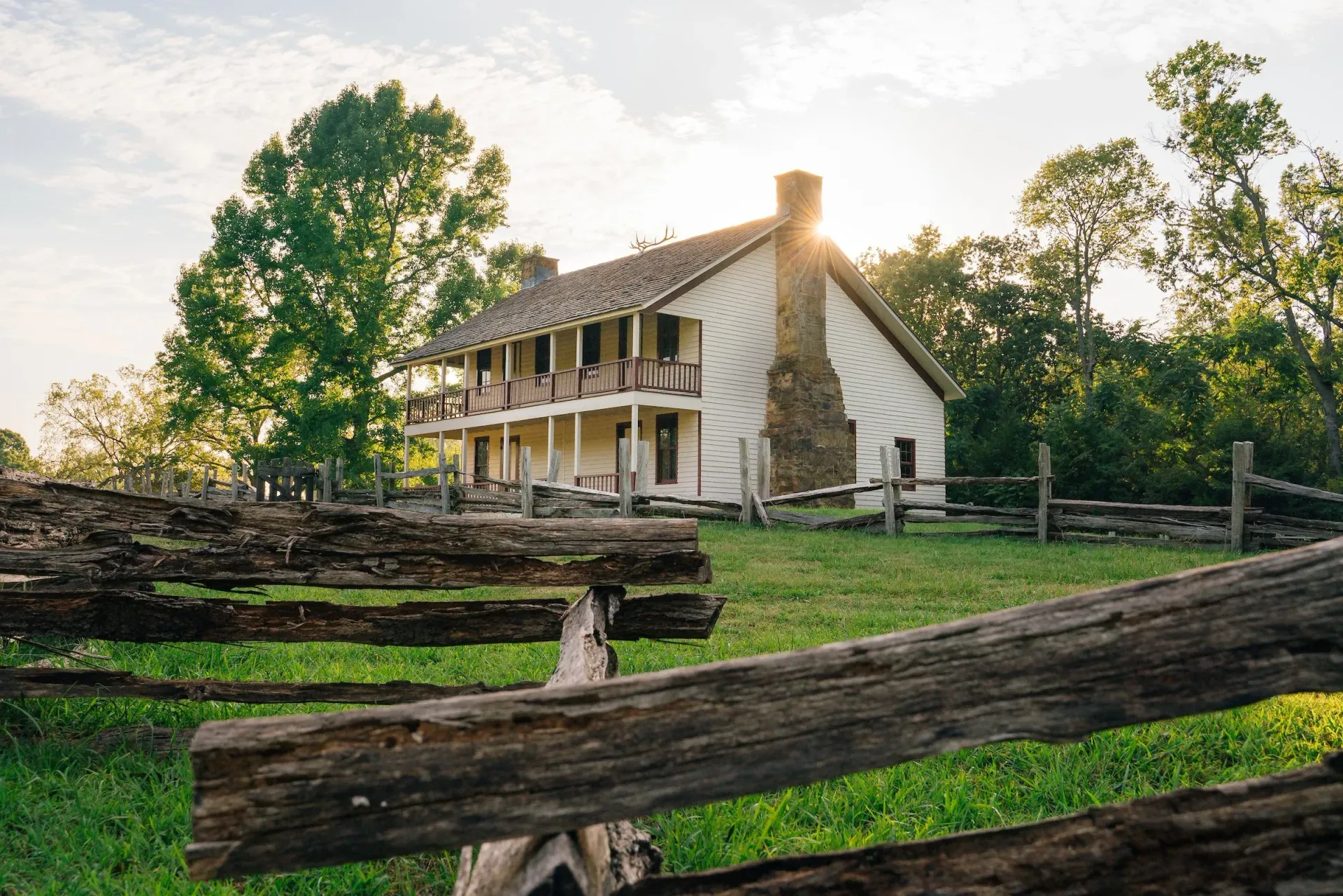Rustic farmhouse with white siding and wooden fence in a sunlit green field at sunset