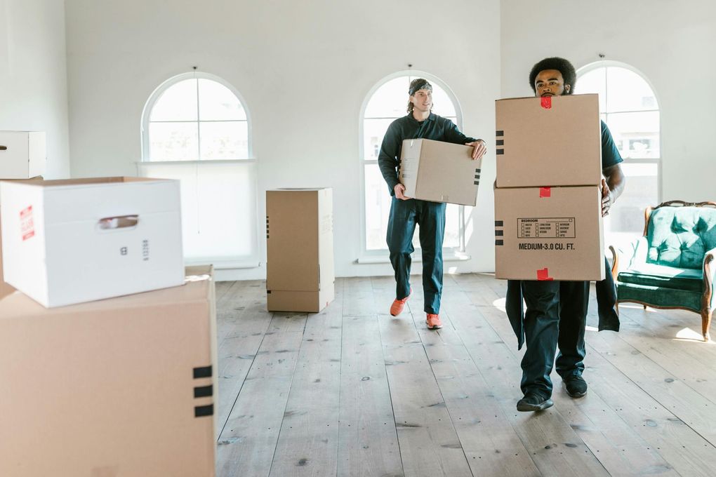 Two people carrying cardboard boxes in a bright room with arched windows and hardwood floors