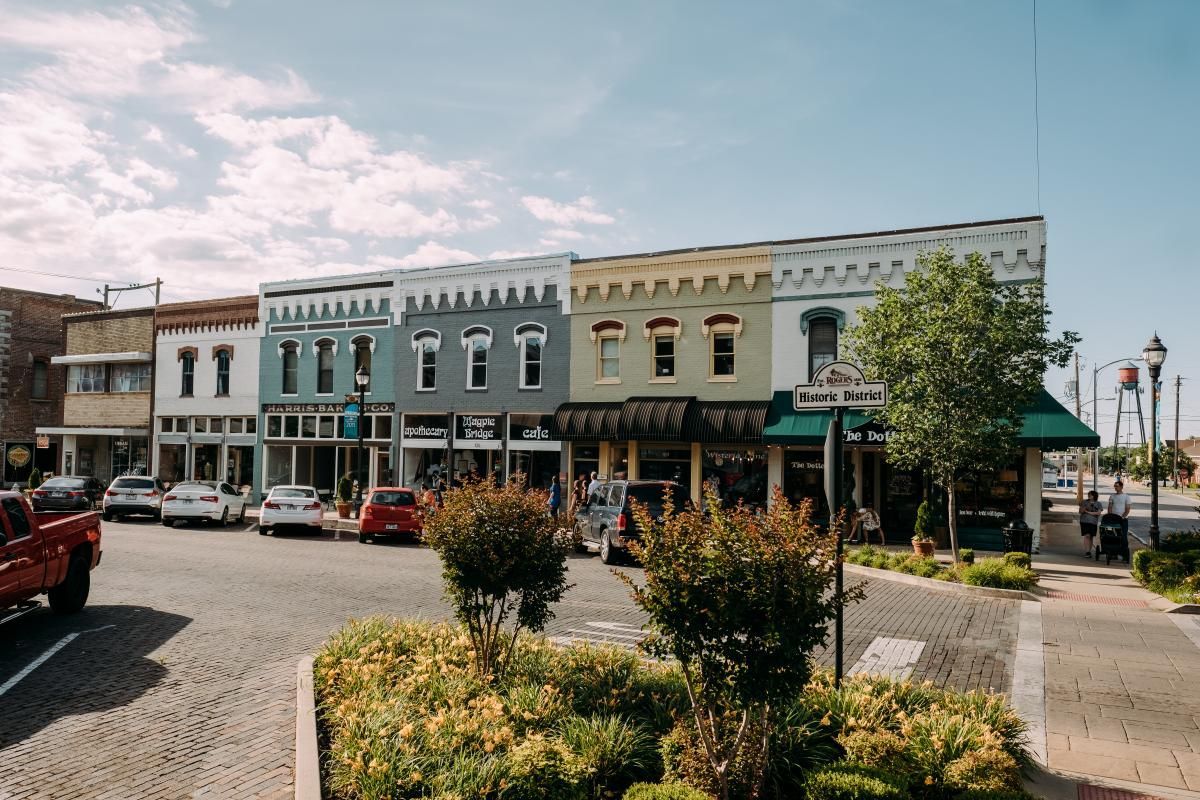 Historic downtown street with colorful storefronts, parked cars, and a tree-lined sidewalk on a sunny day