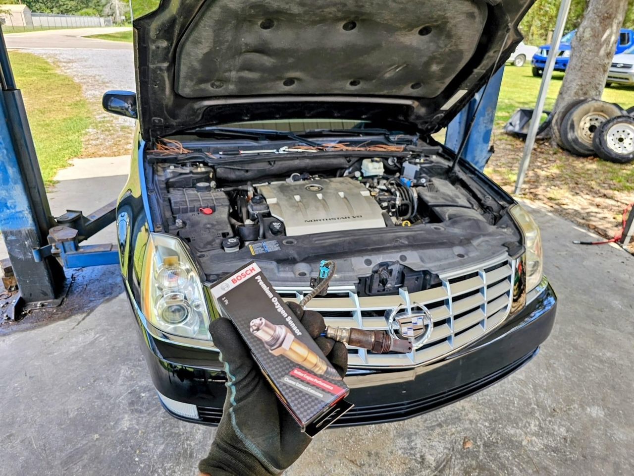 A person is holding a box of spark plugs in front of a car with the hood open.