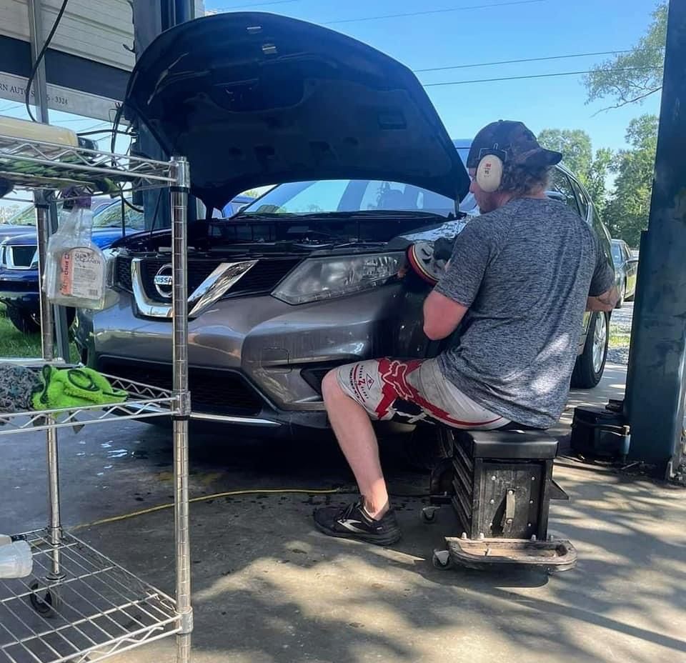 A man is sitting on a stool working on a car with the hood open.