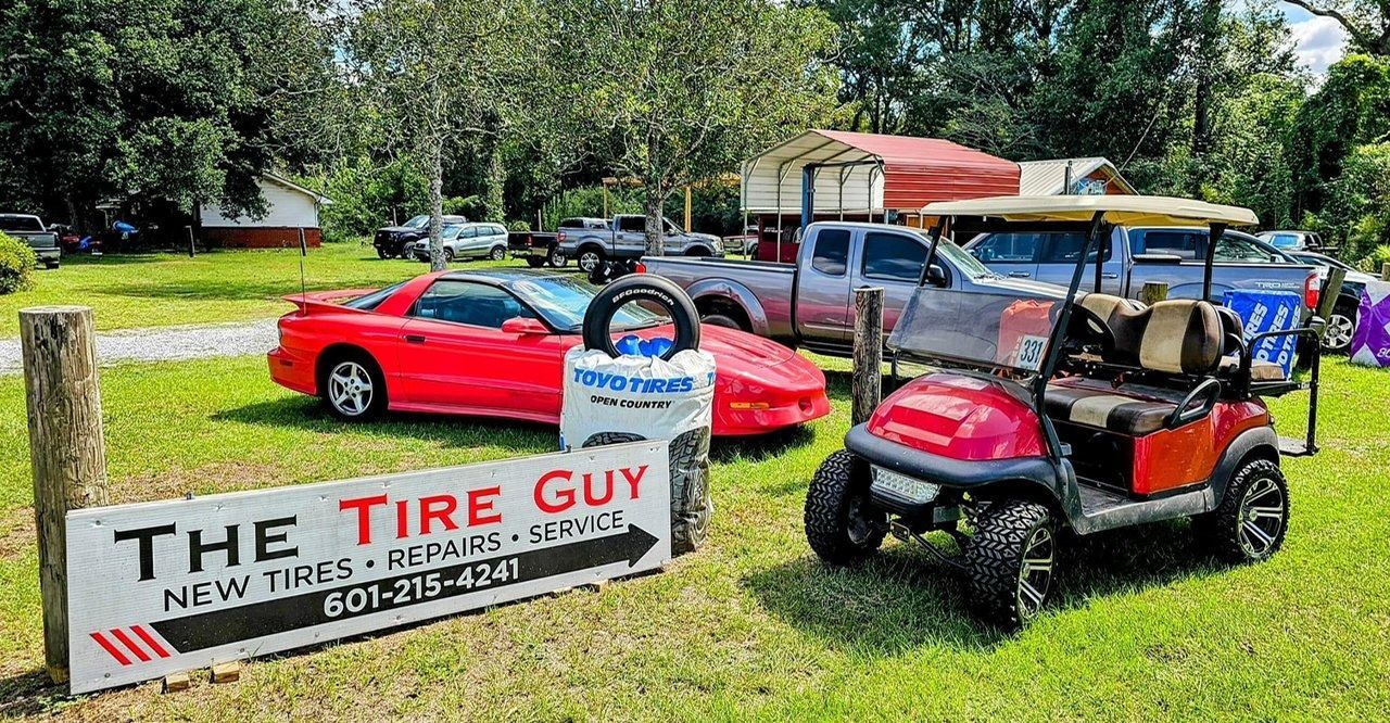A red car and a golf cart are parked in front of a sign for the tire guy.