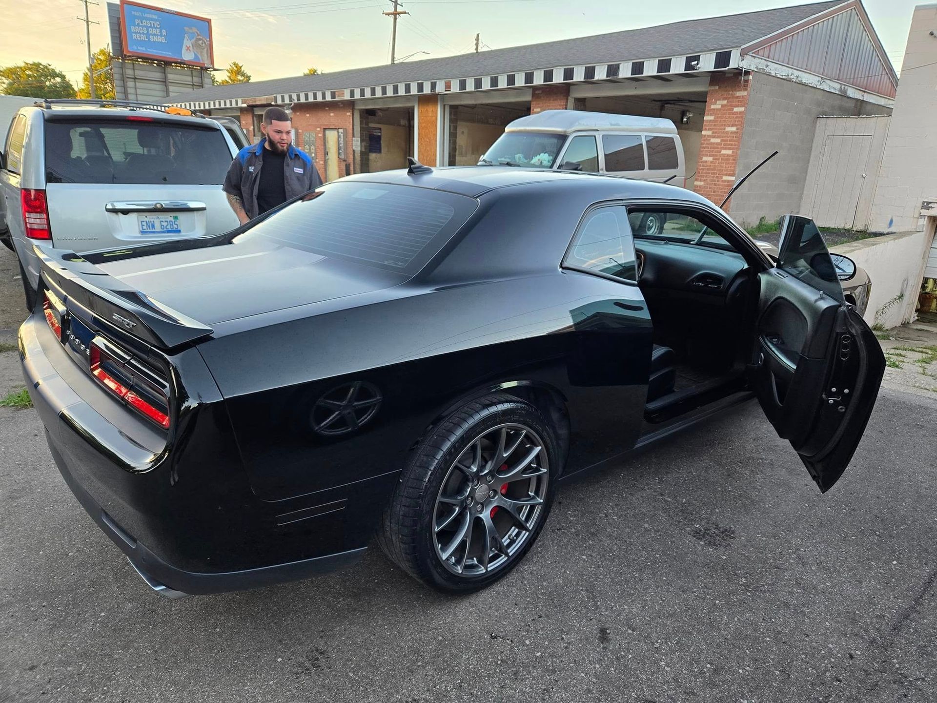 Black Dodge Challenger with open doors, parked outside a building, with a person standing nearby.