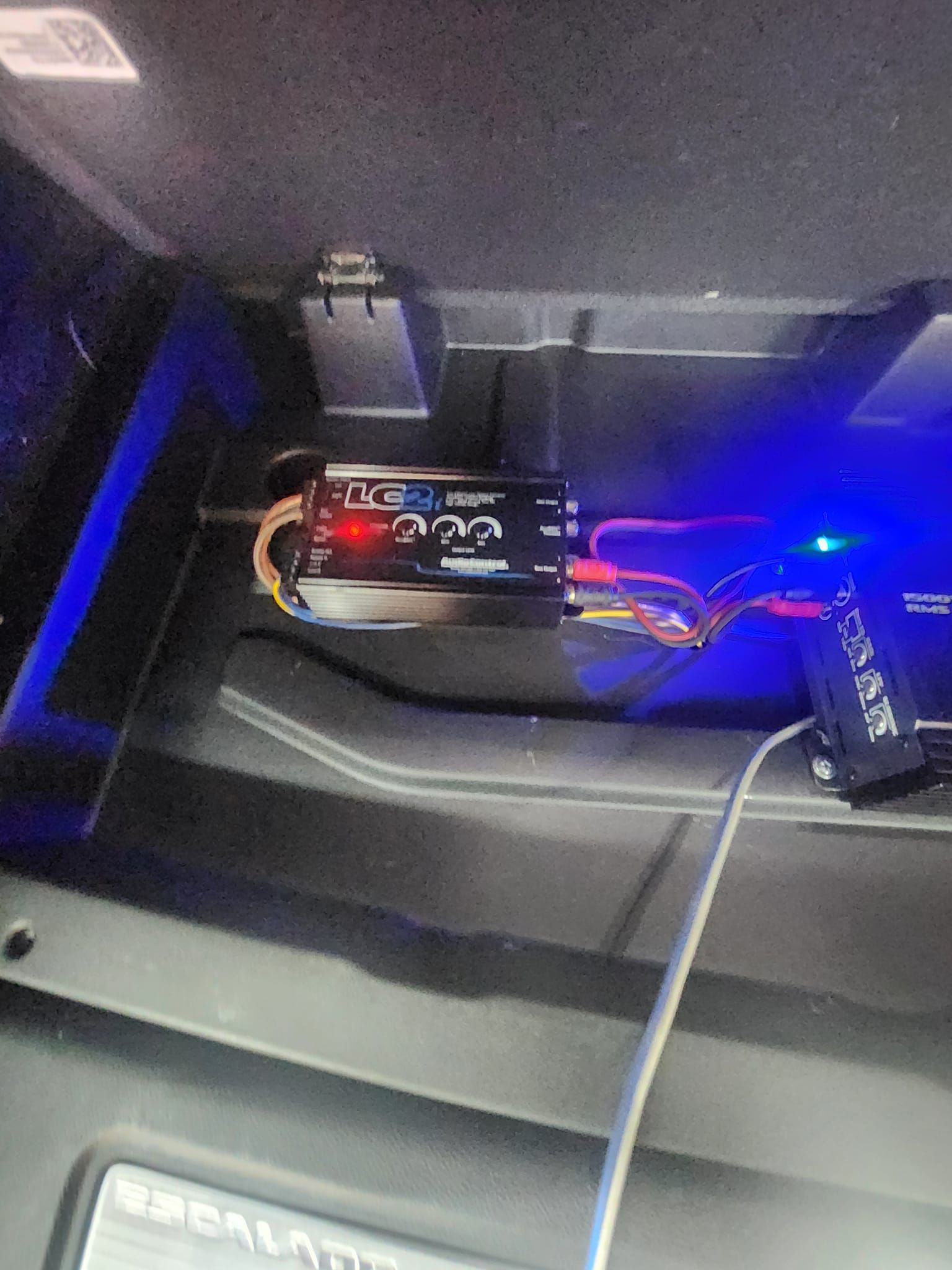 Car audio amplifier and components, black and silver, with blue LED lights, installed in vehicle.