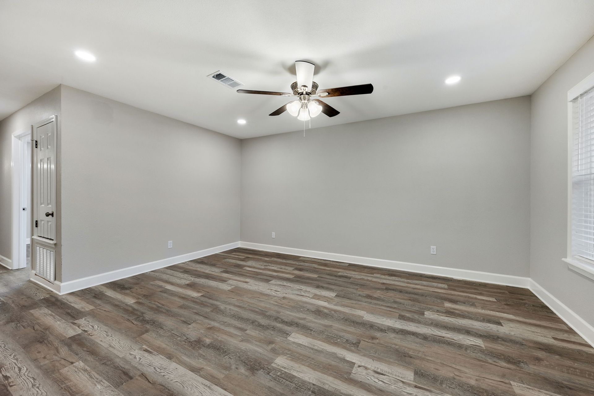 An empty room with light gray walls, brown wood-look flooring, a ceiling fan, and white window blinds.