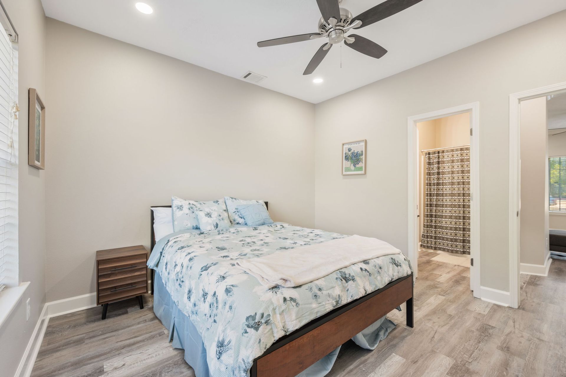 A bedroom with a queen-sized bed, light gray walls, wood-look flooring, a ceiling fan, and an attached bathroom.