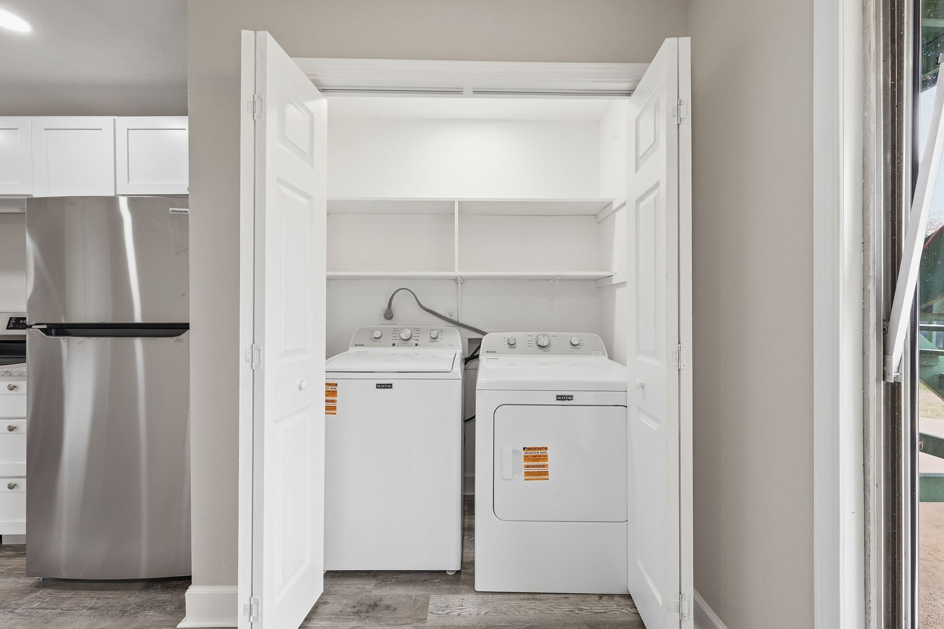 A utility closet containing a white washing machine and dryer, with shelving above, located next to a refrigerator.