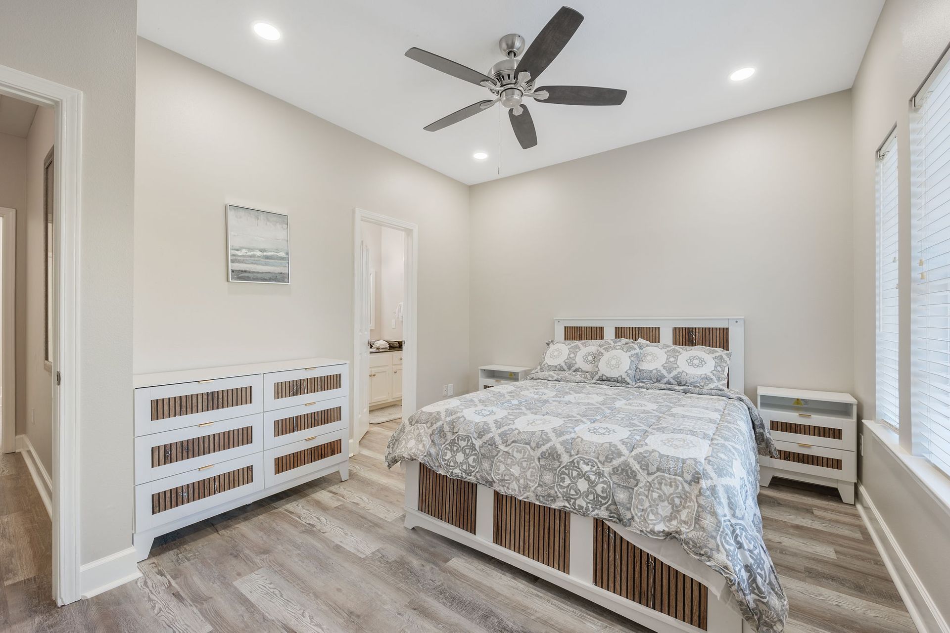 A bright bedroom with a queen bed, white wooden furniture, grey wood-look flooring, and a ceiling fan.