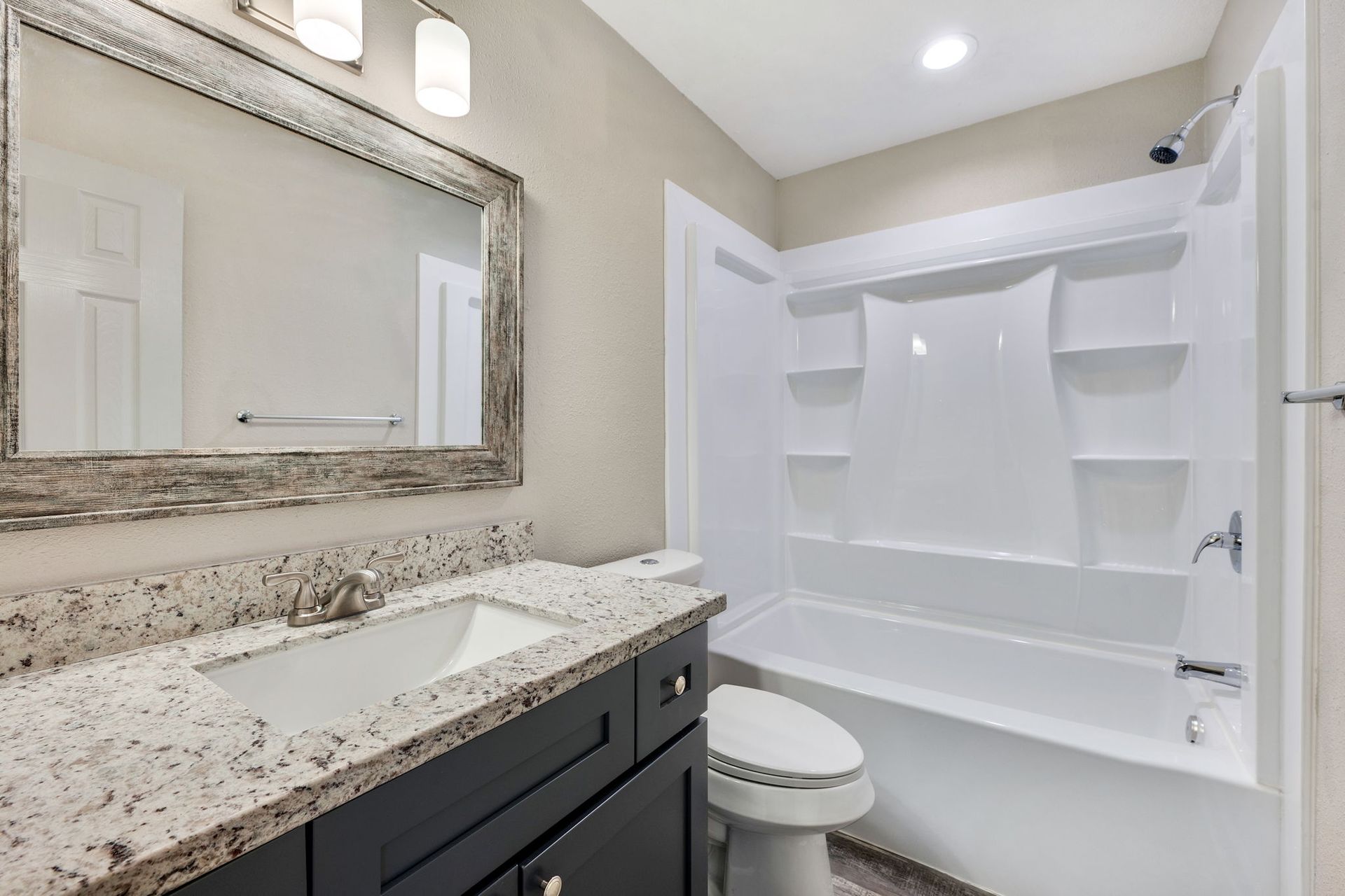 A modern bathroom with a dark vanity, granite countertop, rectangular mirror, and a white tub-shower combo.