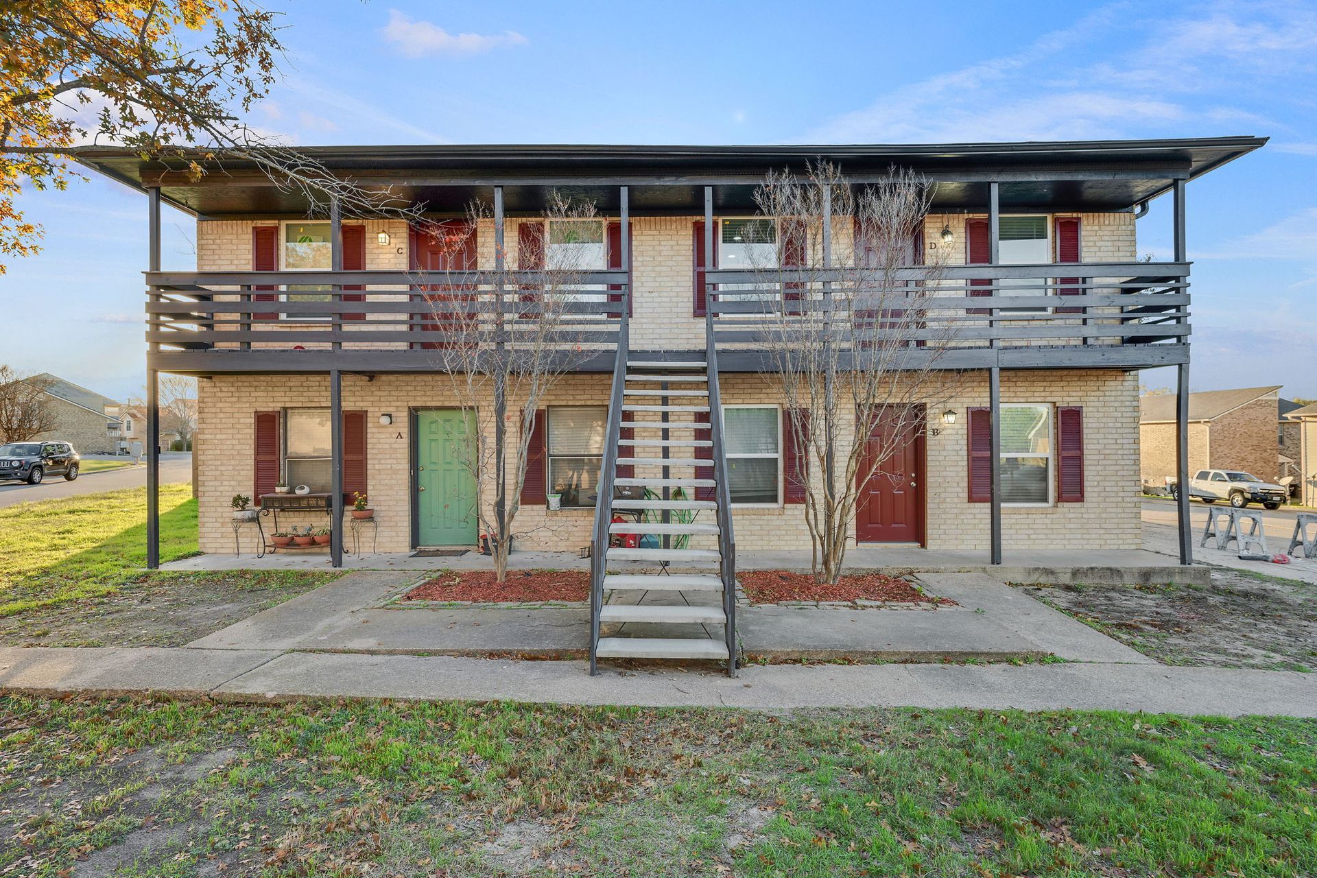 A two-story brick apartment building with a central staircase, balconies, and dark trim under a bright, partly cloudy sky.