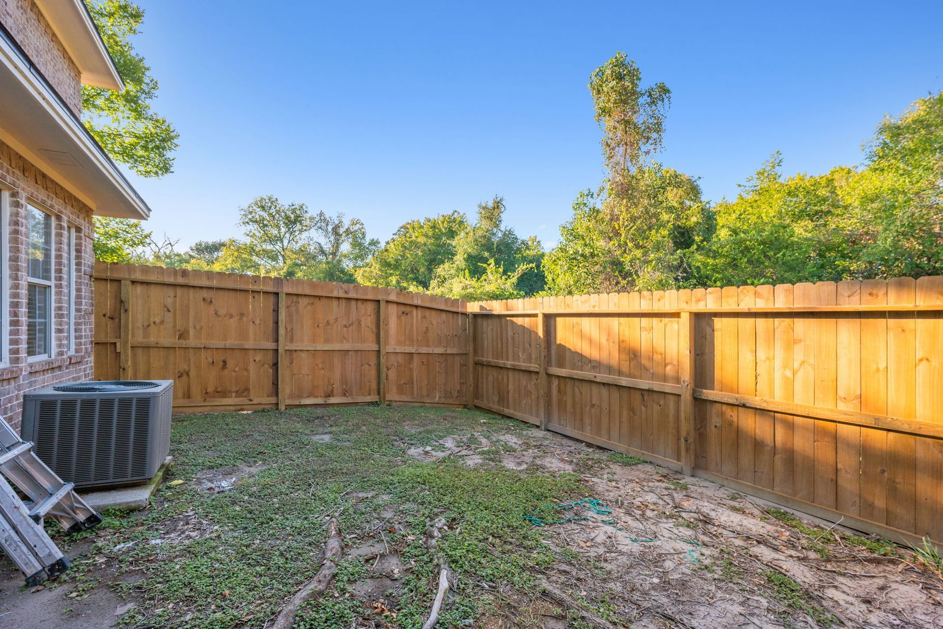 A small backyard space features a brick house corner, a gray HVAC unit, and a tall wood privacy fence against green trees.