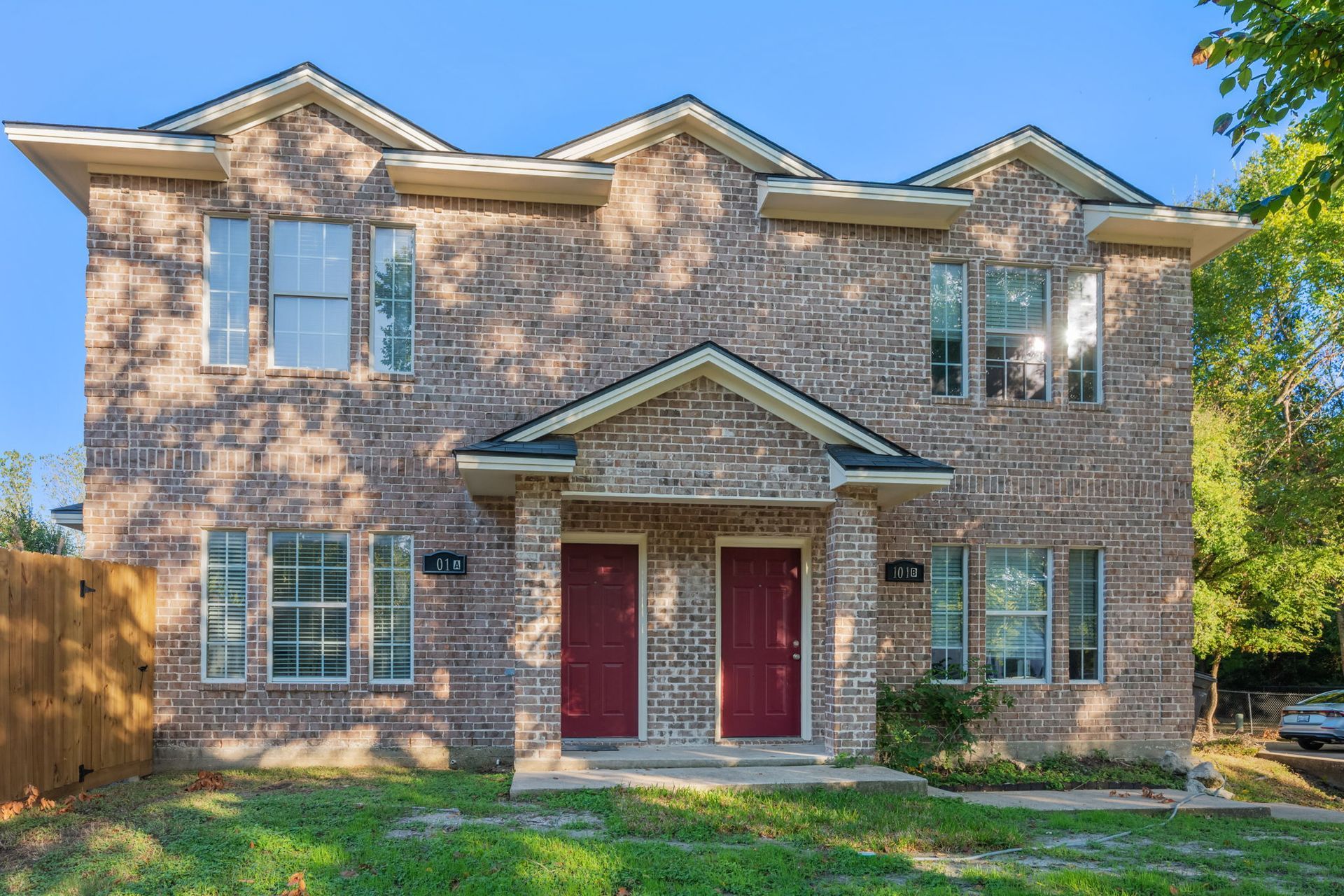 Two-story brick duplex with red front doors and matching brown shingled roofs under a clear blue sky.