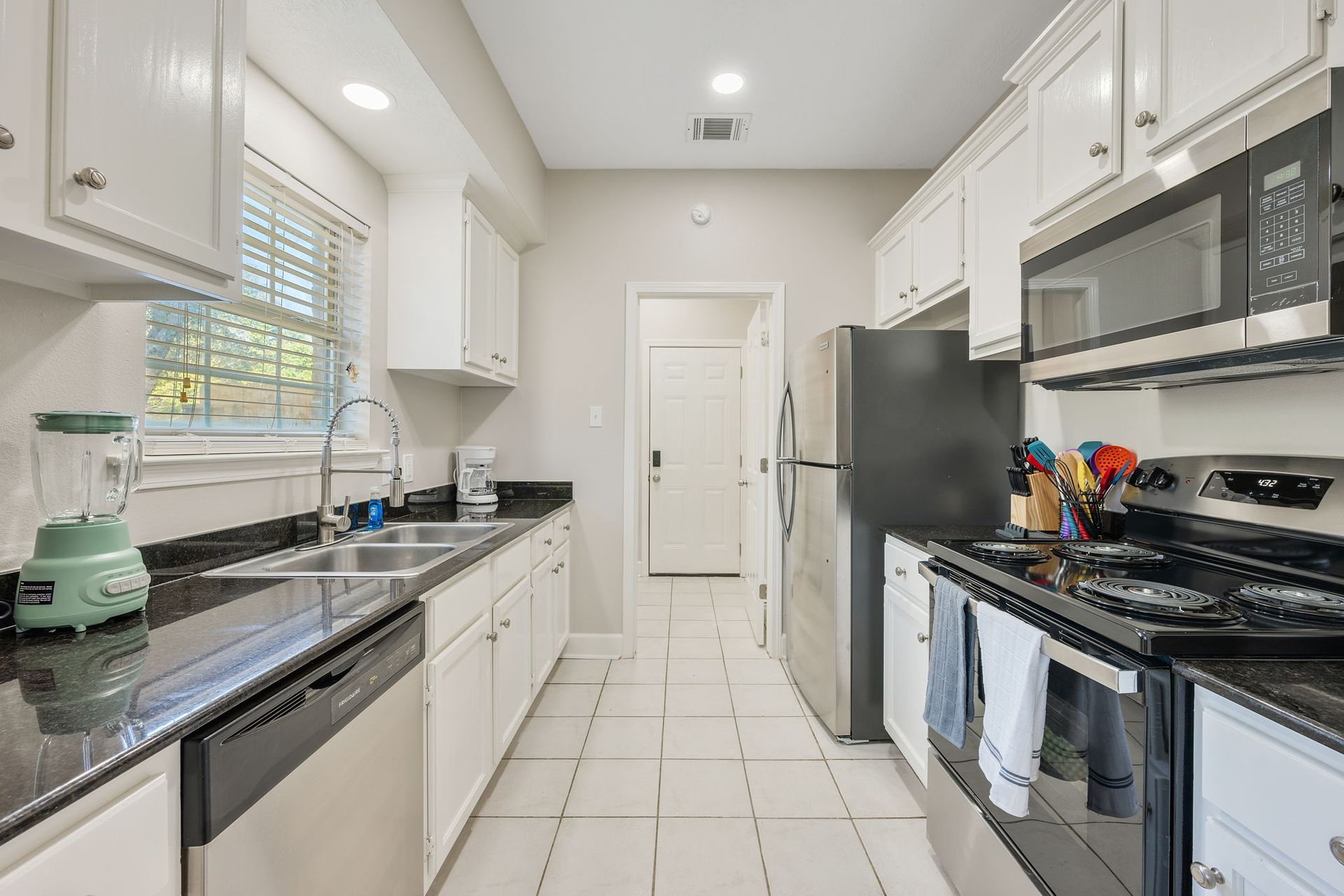 A modern galley kitchen with white cabinets, black countertops, stainless steel appliances, and beige tile flooring.