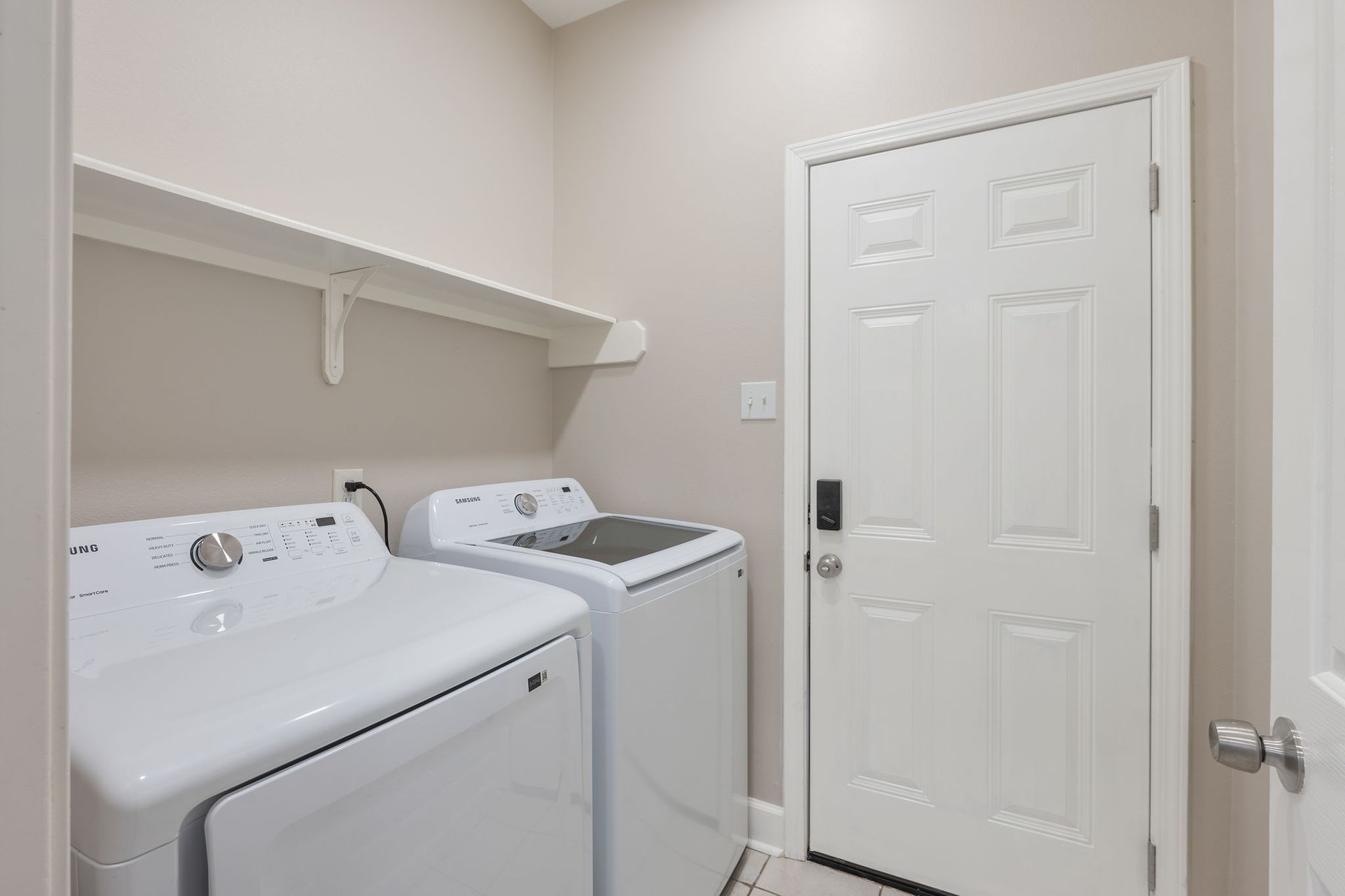 A laundry room with two white machines under a wall-mounted shelf, next to a white door with a digital keypad lock.